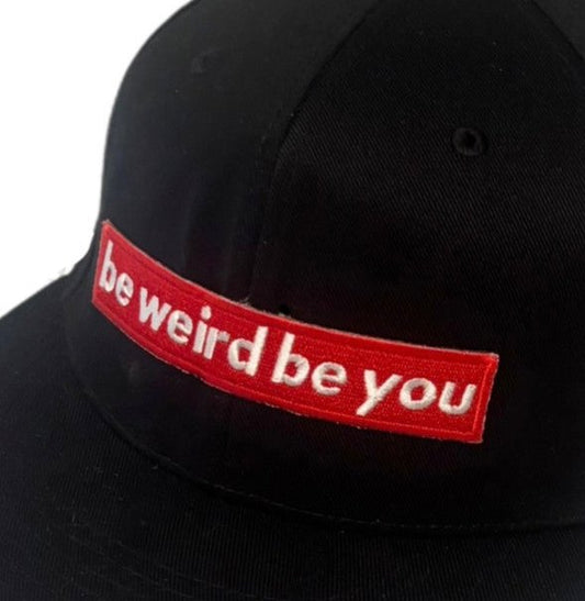 Close-up of red “be weird be you” embroidered patch on a black flat brim hat