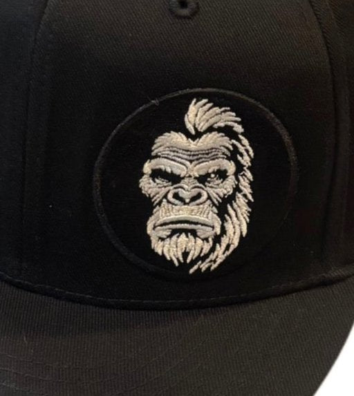 Close-up of embroidered Bigfoot face patch on a black flat brim hat