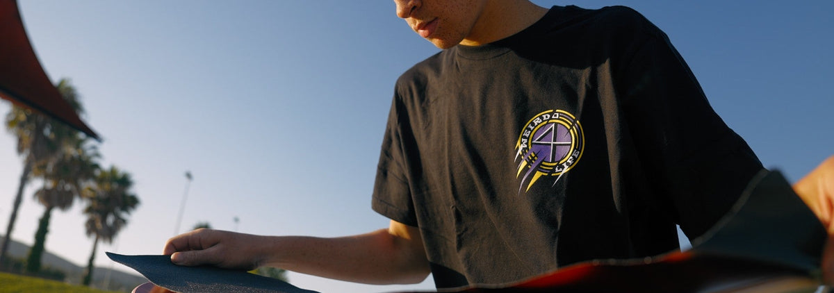 A young man with curly blonde hair sits outdoors in a skatepark, looking down at a skateboard resting on his lap. He is wearing a black t-shirt with a circular graphic logo on the left chest and black pants.