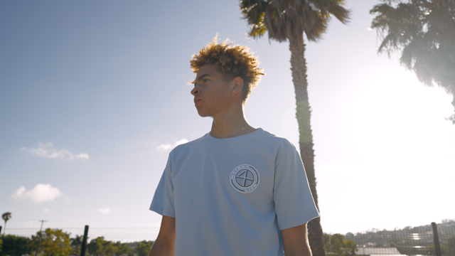A young man with a light blue Weirdo 4 Life T-shirt and palm trees in the background 