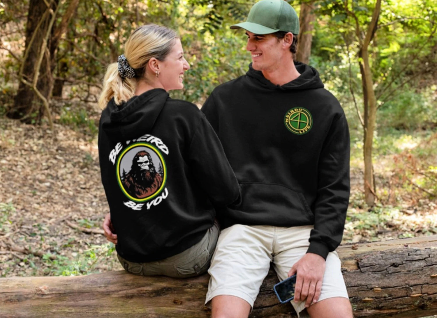 a man and a woman in a forest, sitting on a tree. the man is wearing a green hat and weirdo 4 life hoodie. the woman is wearing a scrunchie in her hair and a weirdo 4 life hoodie.